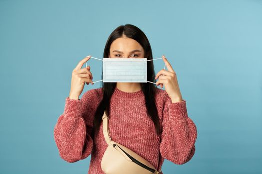 Waist up of pretty lady posing in studio and holding protective face mask, isolated on blue background. Copy space. Quarantine, coronavirus concept