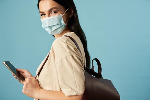 Happy woman holding smartphone while posing in studio with backpack, isolated on blue background. Copy space. Quarantine, epidemic concept