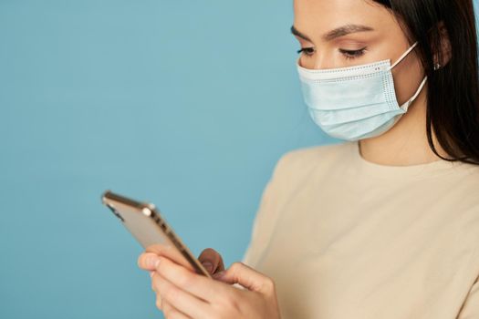 Close up of pretty female wearing protective mask and typing message on mobile phone, isolated on blue background. Copy space. Quarantine, coronavirus concept