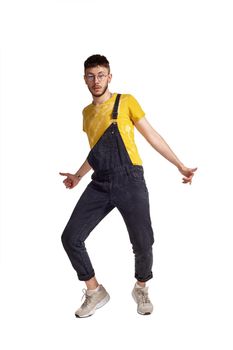 Full-length portrait of a fasionable person in glasses, black jumpsuit, yellow t-shirt and gray sneakers fooling around in studio. Indoor photo of a man making dancing elements isolated on white background and looking at the camera. Music and imagination.