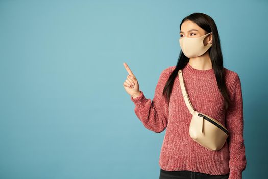 Waist up of beautiful brunette posing in studio and pointing her finger at copy space, isolated on blue background. Quarantine, coronavirus concept