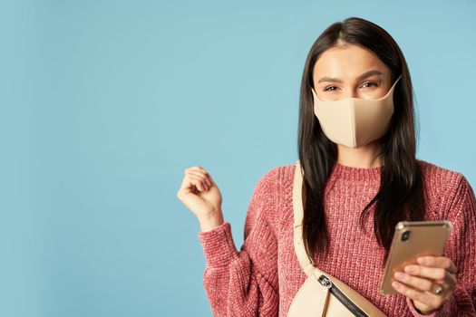 Happy young woman in anti virus face mask holding smartphone while posing in studio, isolated on blue background. Copy space. Concept of travel, coronavirus