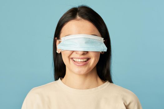 Happy woman in a beige t-shirt posing with a protective mask on her eyes on a blue background. Copy space. Quarantine, epidemic concept