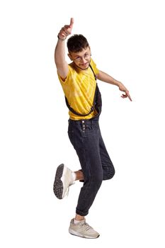 Full-length portrait of an athletic guy in glasses, black jumpsuit, yellow t-shirt and gray sneakers fooling around in studio. Indoor photo of a man making dancing elements raising his index finger isolated on white background. Music and imagination.