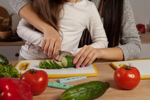 Close-up shot of a happy loving family cooking together. Mom and her kid are making a vegetable salad and cutting a cucumber at the kitchen. Homemade food and little helper.