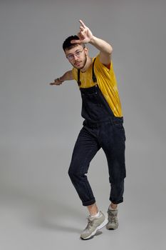 Full-length portrait of a stylish person in glasses, black jumpsuit, yellow t-shirt and gray sneakers fooling around in studio. Indoor photo of a man dancing raising his hands on a gray background. Music and imagination.