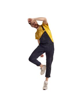 Full-length portrait of a smiling dancer in glasses, black jumpsuit, yellow t-shirt and gray sneakers fooling around in studio. Indoor photo of a man making dancing elements isolated on white background. Music and imagination.