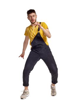 Full-length portrait of a creative person in glasses, black jumpsuit, yellow t-shirt and gray sneakers fooling around in studio. Indoor photo of a man making dancing elements isolated on white background and looking at the camera. Music and imagination.