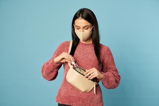 Beautiful brunette in a protective face mask posing in the studio and holding an antiseptic on blue background. Copy space. Quarantine, coronavirus concept