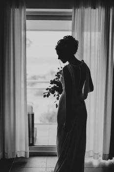 Beautiful bride in a dress with a bouquet of flowers stands, turning around and looking at the floor in the bedroom against the background of the window. Black and white photo. High quality photo