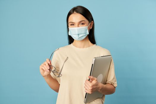 Portrait of pretty young lady using laptop while holding glasses and posing in studio, isolated on blue background. Copy space. Quarantine, epidemic concept