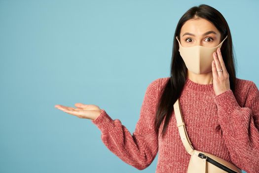 Shocked woman wearing protective face mask and posing in studio while pointing with palm at copy space. Quarantine, coronavirus concept