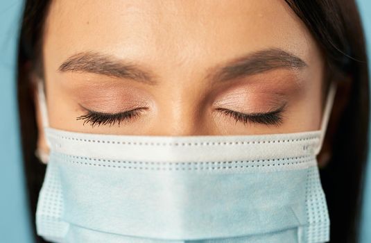 Close up of beautiful Caucasian young woman with closed eyes with disposable face mask, isolated on blue background. Quarantine, coronavirus concept