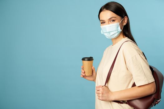 Pretty young woman in face mask holding backpack and coffee in studio, isolated on blue background. Copy space. Quarantine, epidemic concept