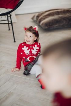 Stock photo portrait of adorable girl in red dress with festive print holding beautifully wrapped golden present in hands while sitting on the floor next to decorated Christmas treee with garland..