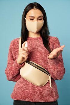 Young woman in anti viral face mask using hand sanitizer spray on blue background in studio. Quarantine, coronavirus concept