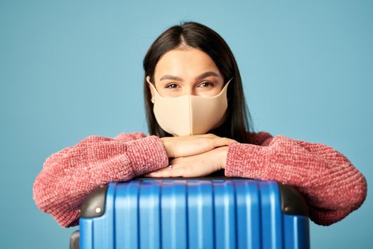 Portrait of beautiful woman posing with suitcase and wearing face mask, isolated on blue background. Copy space. Concept of travel, coronavirus