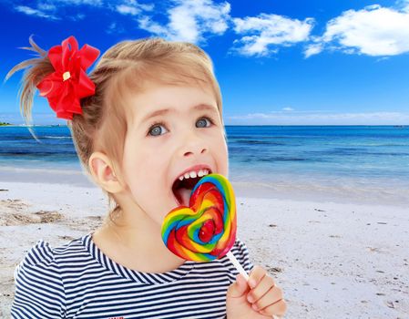 Cute little blonde girl with a red bow on her head, with pleasure licking colorful candy on a stick. Visible language which was painted in a candy color. Close-up.