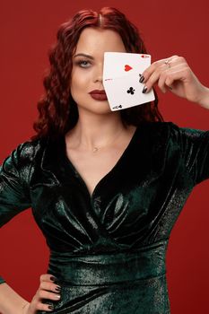 Young sexy woman with a red curly hair, have brought playing cards to her face and looking at the camera, on a red background. Poker
