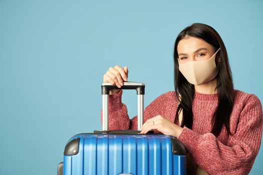 Young woman in a sweater and protective mask holding a suitcase on blue background. Copy space. Concept of travel, coronavirus