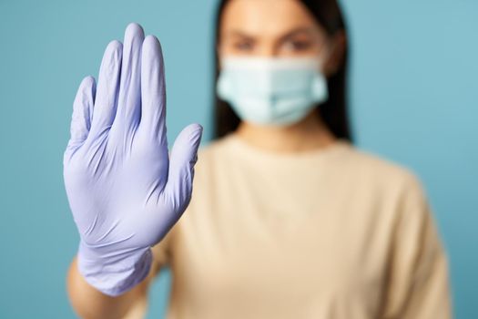 Young woman in protective mask from viruses showing stop sign with gloved hand on blue background. Copy space. Quarantine, epidemic concept