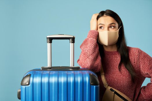 Young woman in protective mask near luggage while waiting for travel, isolated on blue background. Copy space. Concept of travel, coronavirus