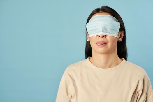 Waist up of woman in a beige t-shirt posing with a protective mask on her eyes on a blue background. Copy space. Quarantine, epidemic concept