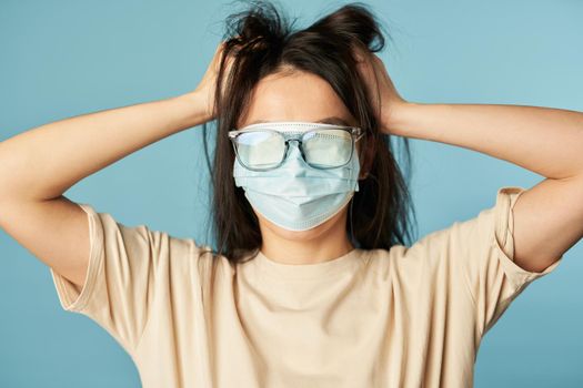 Woman with tousled hair posing with a protective mask and glasses on her eyes on a blue background. Copy space. Quarantine, epidemic concept