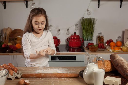 Pretty little brunette child dressed in a white blouse is kneading a dough for baking bread and looking at it at kitchen against a white wall with shelves and bulbs on it. Happy childhood concept.