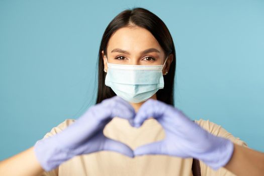 Waist up of pretty young woman in protective face mask posing in studio and showing heart symbol with hands. Copy space. Quarantine, epidemic concept