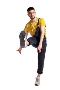 Full-length portrait of fasionable male in glasses, black jumpsuit, yellow t-shirt and gray sneakers fooling around in studio. Indoor photo of a man making dancing elements rsising his leg isolated on white background. Music and imagination.