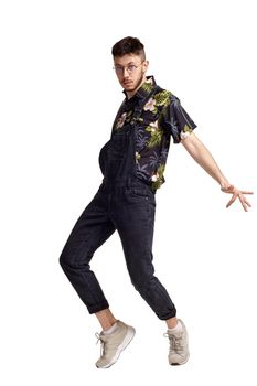 Full-length portrait of a handsome person in glasses, black jumpsuit, colorful t-shirt and gray sneakers fooling around in studio. Indoor photo of a man dancing sideways isolated on white background. Music and imagination.