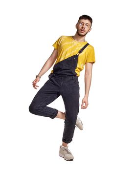 Full-length portrait of an athletic person in glasses, black jumpsuit, yellow t-shirt and gray sneakers fooling around in studio. Indoor photo of a man making dancing elements isolated on white background. Music and imagination.