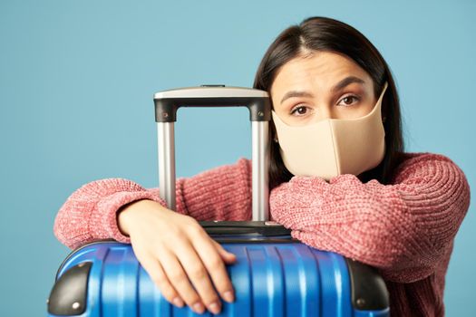 Close up of young woman wearing a protective face mask and leaning on suitcase on blue background. Copy space. Concept of travel, coronavirus