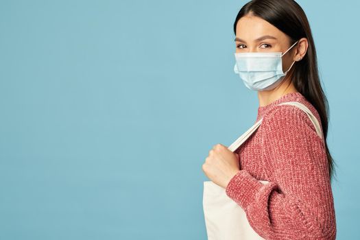 Side view of beautiful young lady posing in studio while wearing face mask and holding shopping bag. Copy space. Quarantine, coronavirus concept