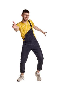 Full-length portrait of a funny person in glasses, black jumpsuit, yellow t-shirt and gray sneakers fooling around in studio. Indoor photo of a smiling man making dancing elements isolated on white background. Music and imagination.