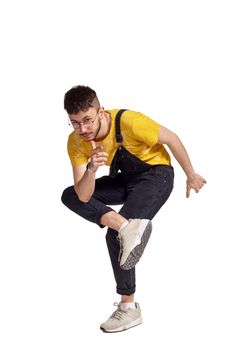 Full-length portrait of a good-looking guy in glasses, black jumpsuit, yellow t-shirt and gray sneakers fooling around in studio. Indoor photo of a man making dancing elements isolated on white background and looking down. Music and imagination.