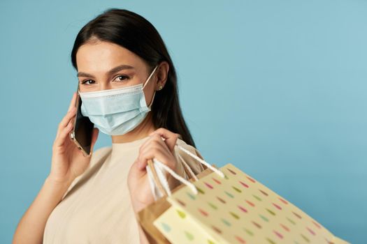 Waist up of pretty female in disposable face mask holding shopping bags while talking on smartphone. Copy space. Quarantine, coronavirus concept