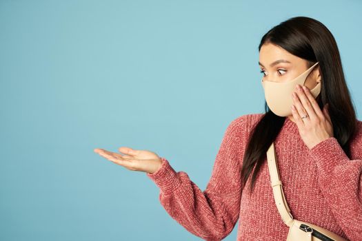 Surprised lady in protective mask touching her cheeks with her hand and looking to the side on blue background. Copy space. Quarantine, coronavirus concept