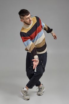 Full-length photo of a handsome male in glasses, black jumpsuit, multi-colored sweater and gray sneakers fooling around in studio. Indoor portrait of a man dancing on a gray background. Music and imagination.