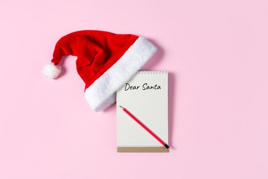 Dear Santa letter concept. Writing in Notepad letter to Santa Claus on pink background with Santa hat .