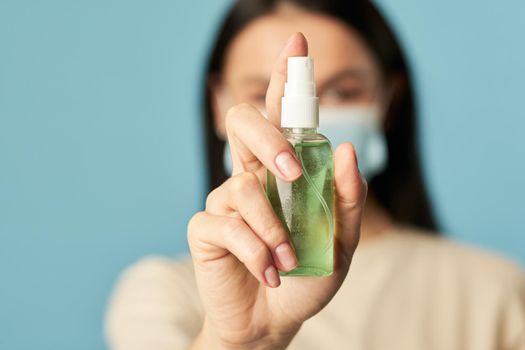 Close up of woman in a protective face mask posing in the studio and holding an antiseptic on blue background. Quarantine, coronavirus concept