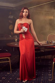 Pretty redheaded lady in a long red dress with an open sexy belly. She is holding two playing cards in her hand and posing standing against a poker table in luxury casino. Passion, chips, alcohol, win, gambling - it is as female entertainment. Smoke background.
