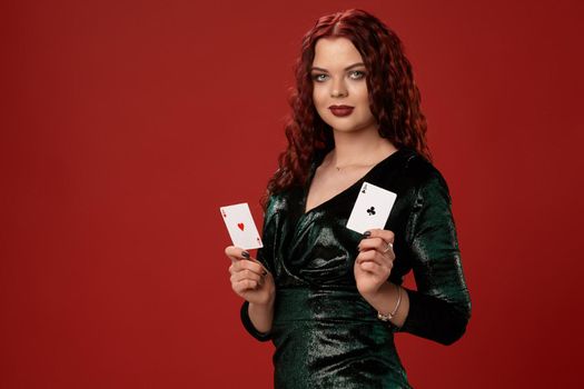 Young sexy woman with a red curly hair, holding aces in her hands and posing sideways, on a red background. Poker
