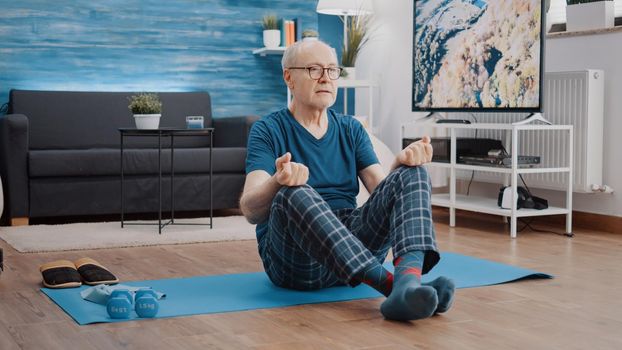 Calm pensioner in lotus position doing meditation to relax on yoga mat. Senior man meditating to practice wellness and relaxation. Aged person doing pilates activity for harmony.