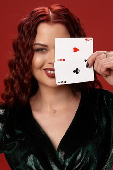Young sexy woman with a red curly hair, have brought playing cards to her face and smiling, on a red background. Poker