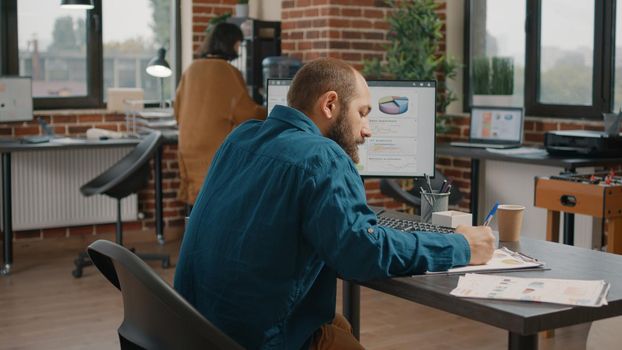 Man taking notes on charts papers and looking at computer screen. Business employee working with data and information on monitor, writing on files to plan project and presentation.