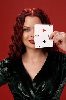 Pretty woman with a red curly hair, have brought playing cards to her face and smiling, on a red background. Poker