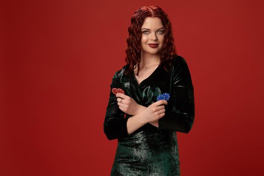 Pretty young redheaded woman is holding some gambling chips in her crossed hands and smiling, on a red background.