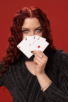 Beautiful redheaded woman in a black knitted dress have covered her face with some playing cards. Casino. Four aces. Studio shot on a red background.